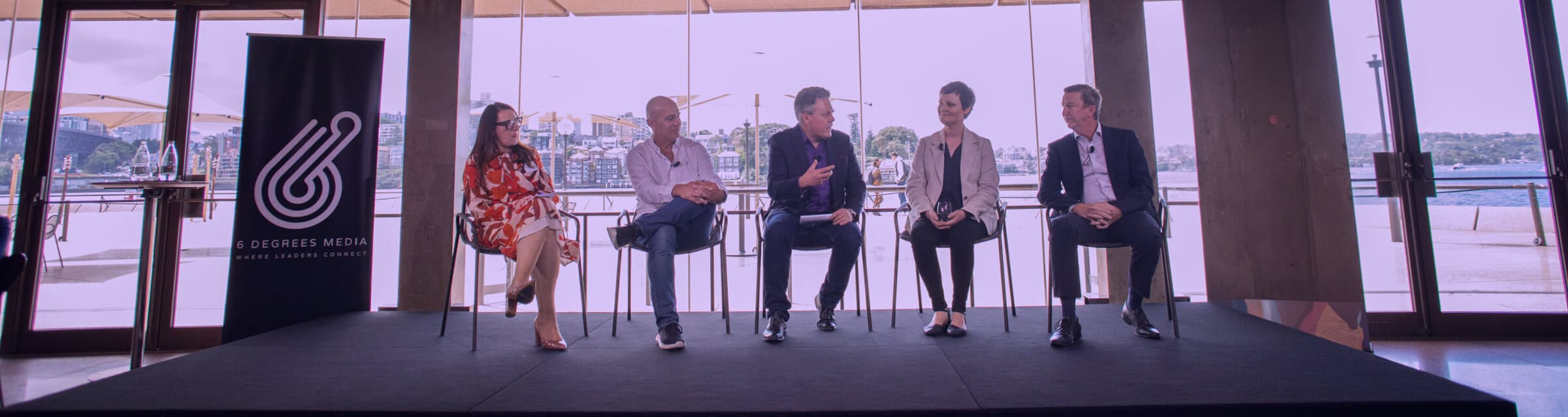 Mark Jones hosting a harbourside panel of five, centre of the row, mid-answer with the harbour behind
