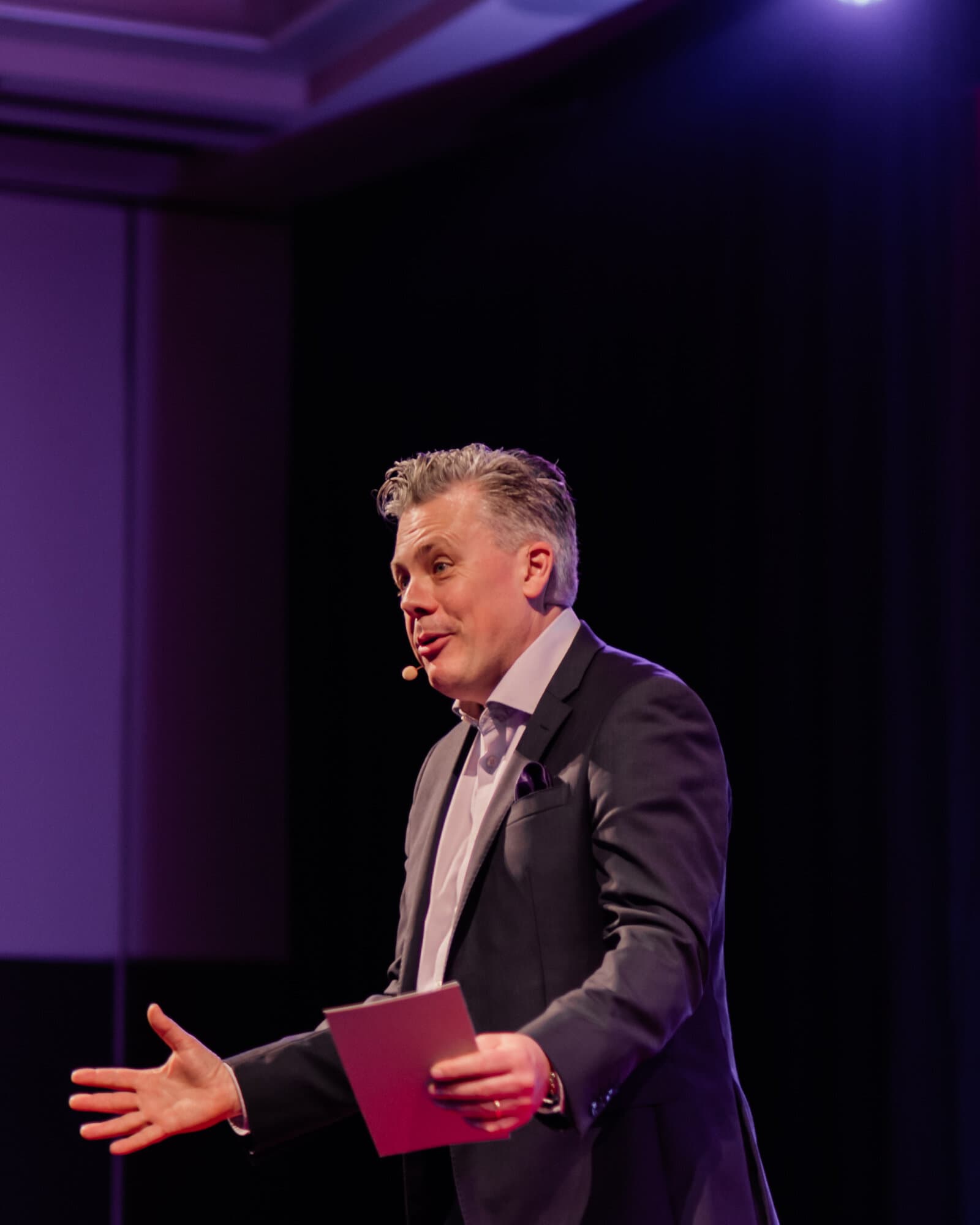 Mark Jones hosting from the stage, host cards in hand, arm open to the room mid-laugh