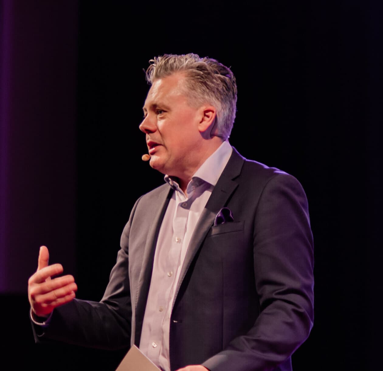 Mark Jones on stage mid-sentence, lit against a violet wall, notes in hand
